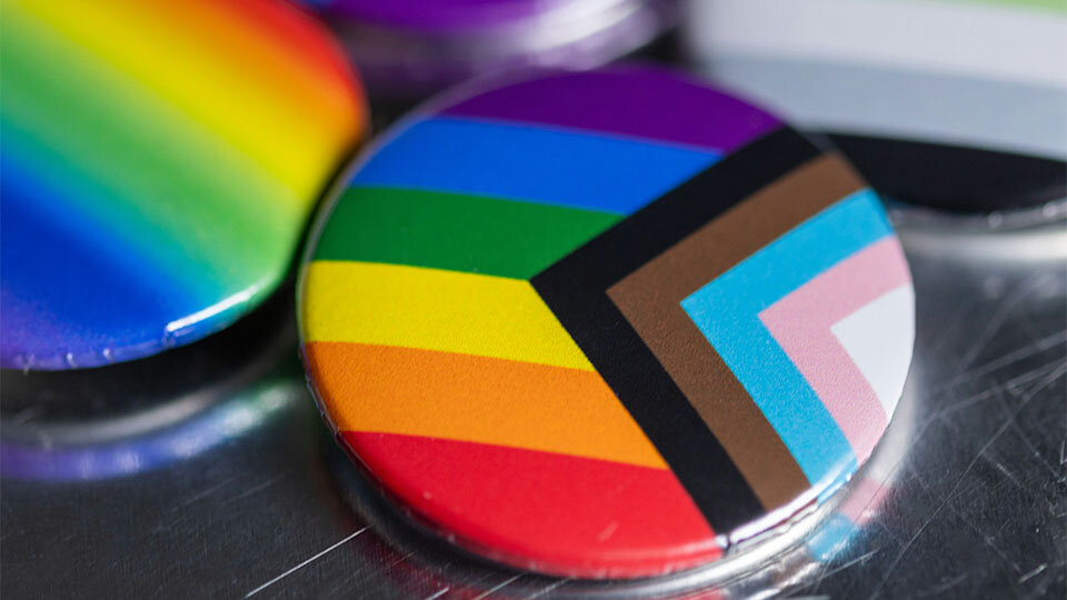 Progress Pride flag pin with vibrant stripes on a reflective surface.