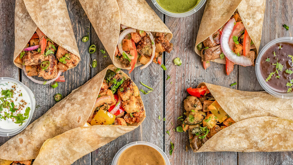 Assorted wraps with colorful vegetables and meat on a wooden table, surrounded by dipping sauces.