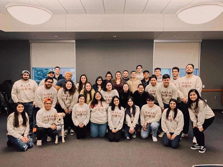 Group of people wearing matching sweaters, posing indoors, with a dog in front. Mexican American Student Association.
